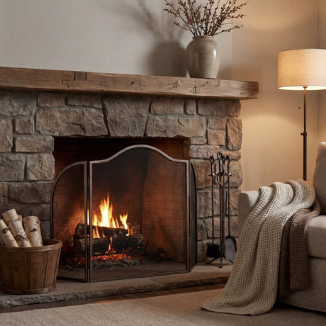 Hill Interiors Arched Three-Panel Fire Screen in silver metal placed before a lit stone fireplace with wooden mantel, log basket and companion set.
