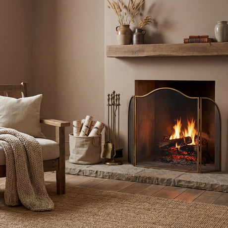 Hill Interiors Large Brass Fire Screen with an arched three-panel mesh design, set before a lit fireplace beside a log basket and companion tool set.