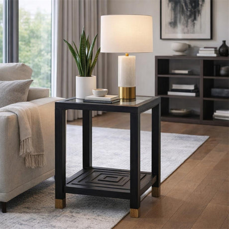 Hill Interiors Harweld wooden side table in matt black with square top, lower shelf and brass-tipped feet, styled with a lamp and plant in a living room.