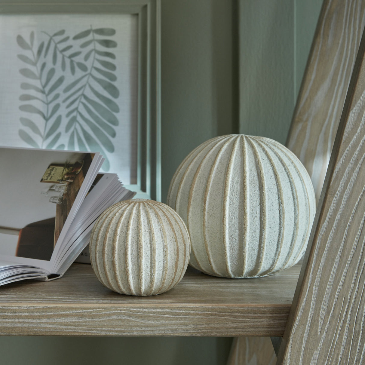 Hill Interiors Medium Decorative Ceramic Ball in white with ribbed spherical form, styled on a wood shelf beside a book and framed leaf print.