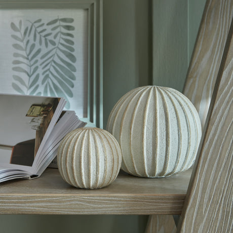 Hill Interiors Medium Decorative Ceramic Ball in white with ribbed spherical form, styled on a wood shelf beside a book and framed leaf print.