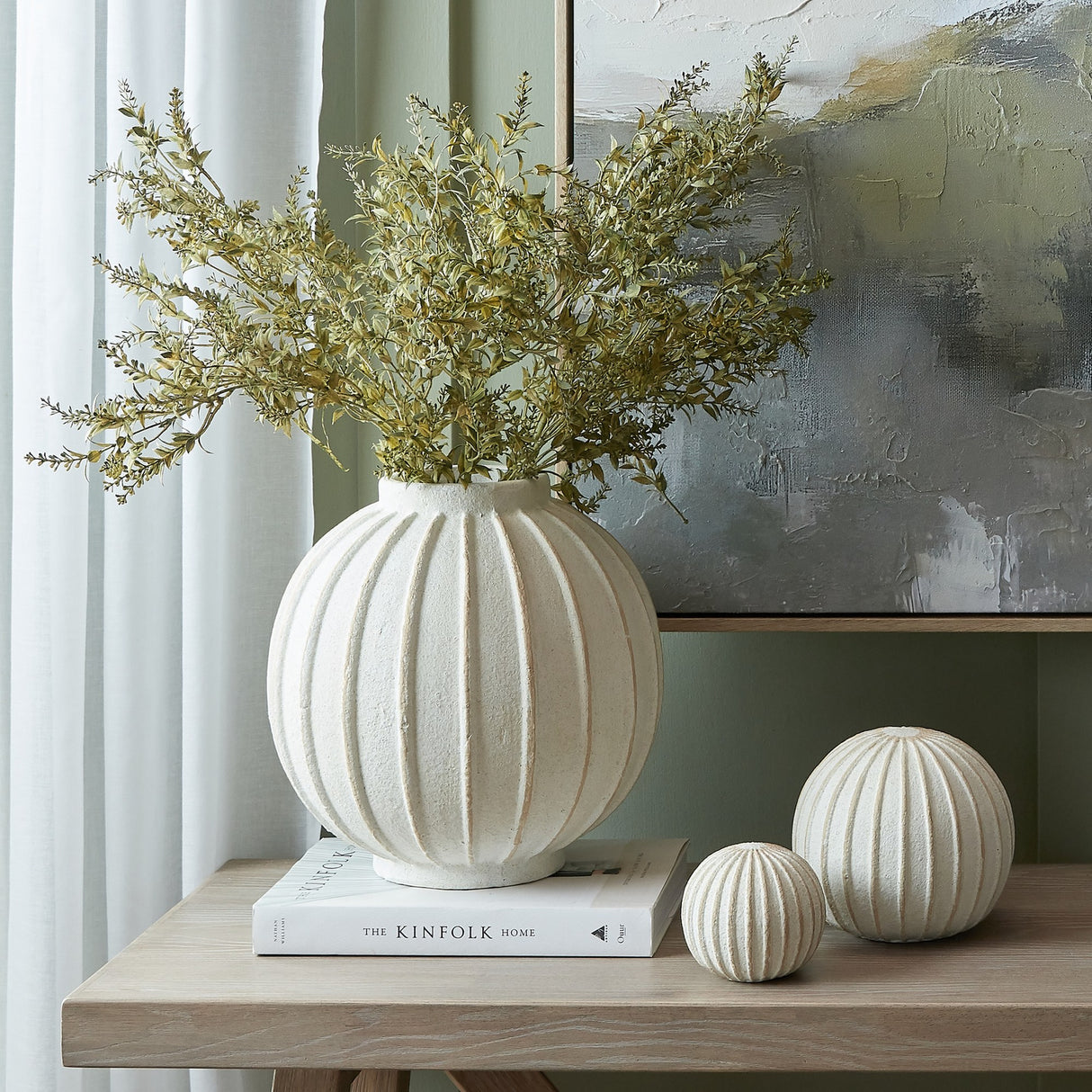 Hill Interiors Large Globe Ceramic Vase in white with ribbed spherical body holding leafy stems, styled on a book beside two smaller ribbed spheres.