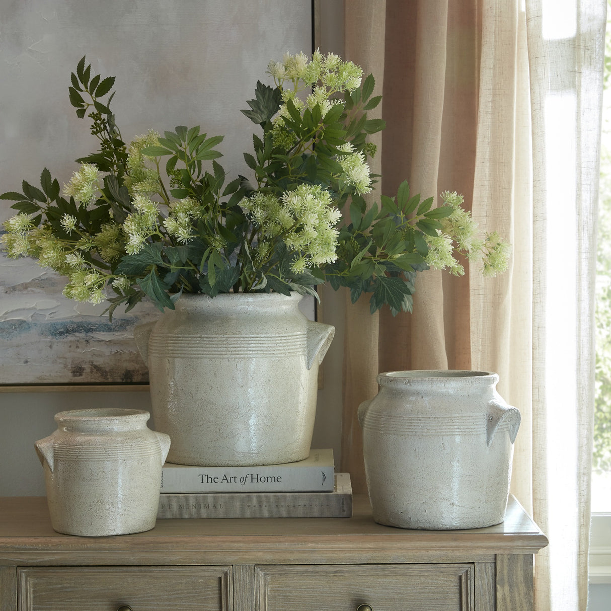 Hill Interiors Medium Ceramic Pot With Handles in white displayed with two smaller matching pots and a leafy white floral arrangement on a wooden sideboard.