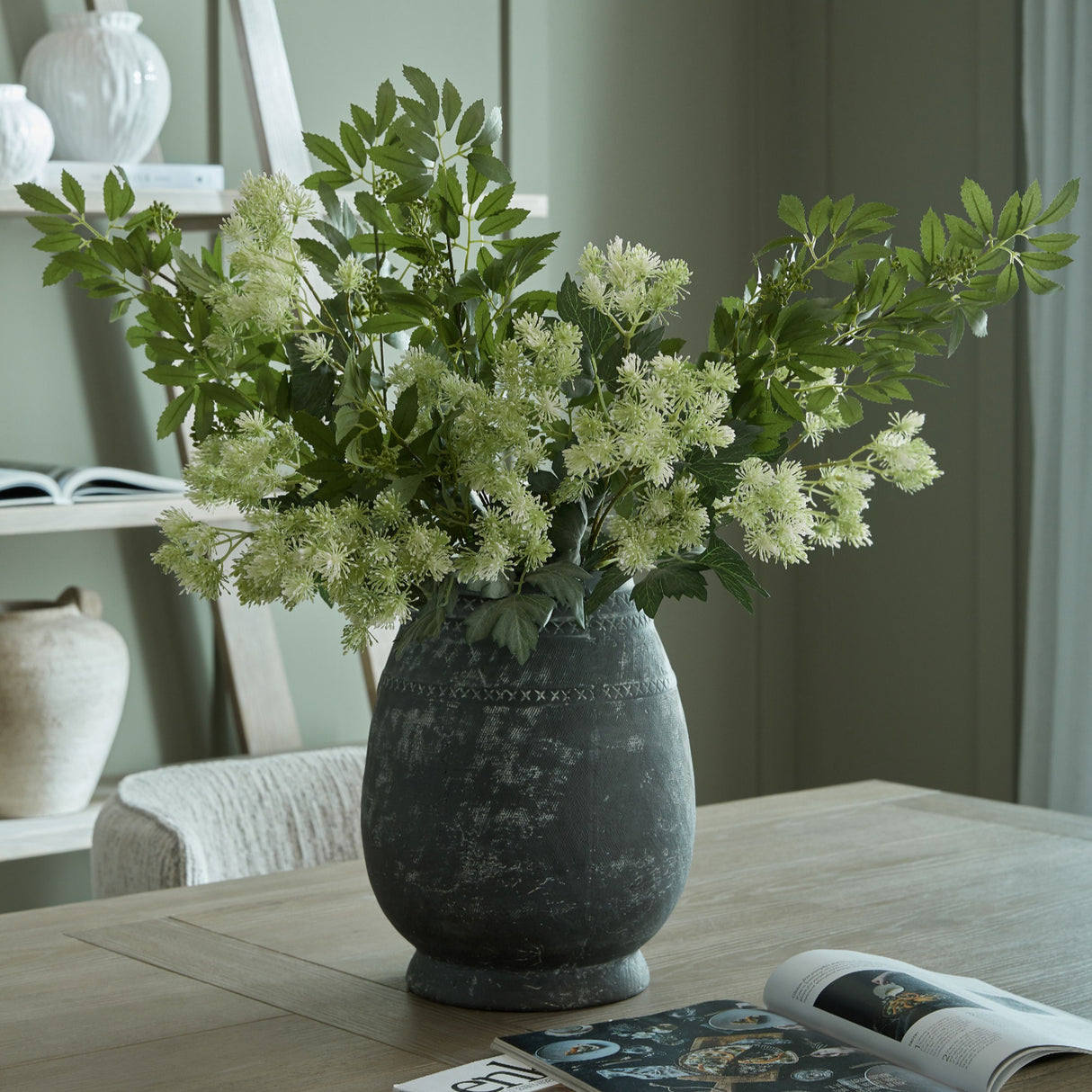 Hill Interiors Etched Tall Ceramic Vase in black with rounded body and textured pattern, holding white flowering stems on a wooden table in a living room.