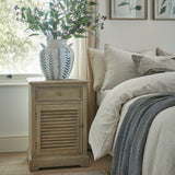 Hill Interiors brown wood bedside cabinet with single drawer over a louvred left-hand door, styled beside a linen-dressed bed with a blue floral vase on top.