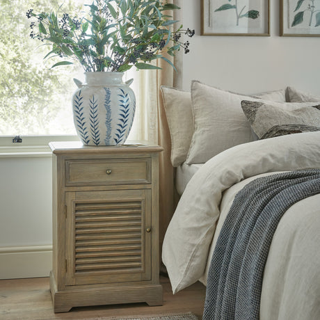 Hill Interiors brown wood bedside cabinet with single drawer over a louvred left-hand door, styled beside a linen-dressed bed with a blue floral vase on top.