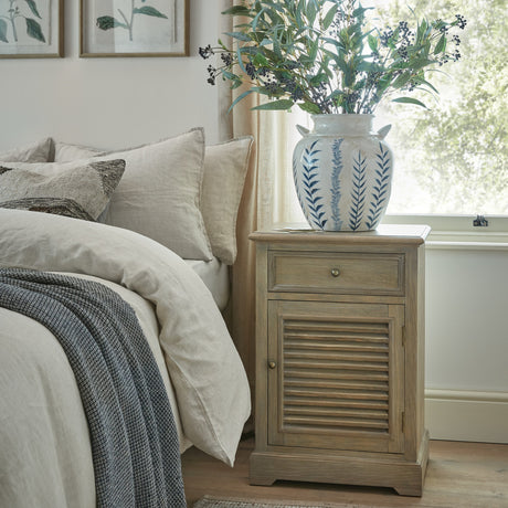 Hill Interiors Wooden Louvred One-Door Chest in brown, used as a bedside cabinet with a blue and white leafy vase, beside a linen-dressed bed.