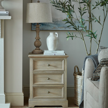 Hill Interiors Wooden Three-Drawer Chest in brown styled in a living room with a lamp, ceramic jug and books on top beside a sofa and olive tree.