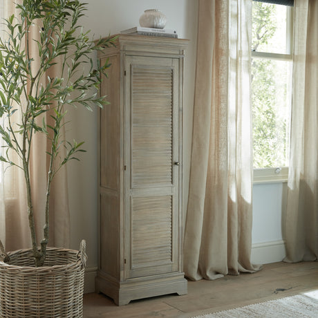 Hill Interiors 1 Door Tall Cabinet in brown wood with louvred door, styled by a window with linen curtains, olive tree and books on top.