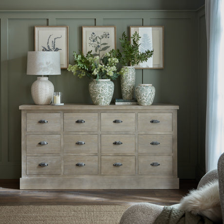 Hill Interiors 12 drawer wooden chest of drawers in warm brown, styled with a lamp, floral vases and framed botanical prints against a sage panelled wall.