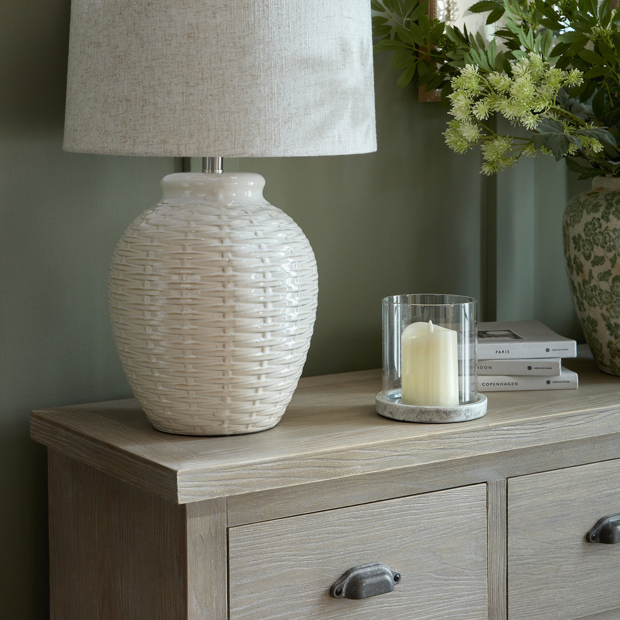 Hill Interiors wooden chest of drawers styled with a woven ceramic lamp, candle in glass holder, books and floral vase against a sage green wall.