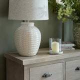 Hill Interiors wooden chest of drawers styled with a woven ceramic lamp, candle in glass holder, books and floral vase against a sage green wall.