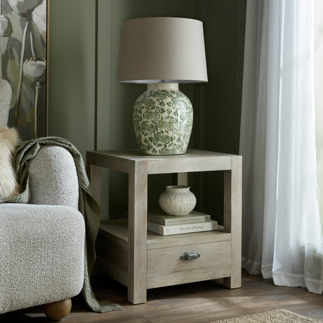 Hill Interiors wooden one-drawer side table in brown with open shelf, styled with a floral ceramic lamp beside a boucle armchair in a green room.