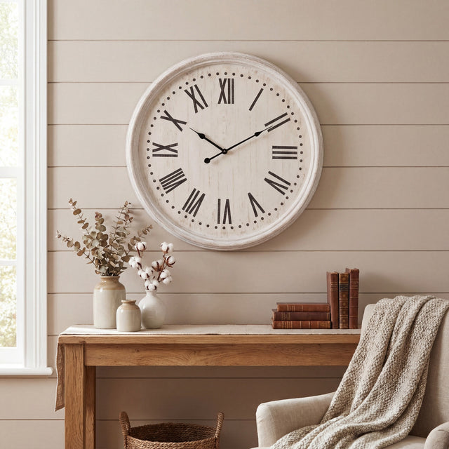 Hill Interiors white wash wooden wall clock with black Roman numerals, mounted above a rustic console table with vases and books in a neutral living room.