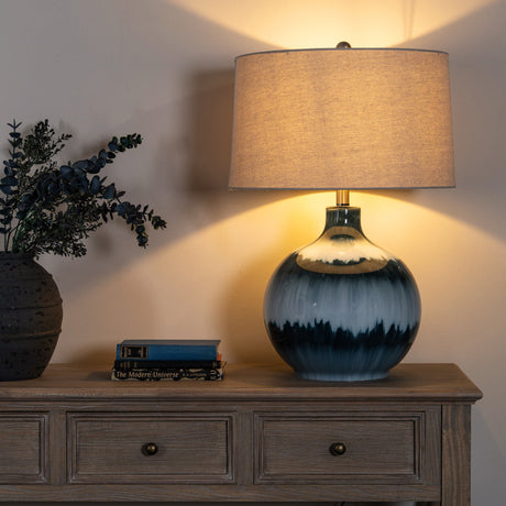 Hill Interiors Glass Table Lamp with rippled blue moulded glass base and natural linen drum shade, lit on a wooden console beside eucalyptus and books.