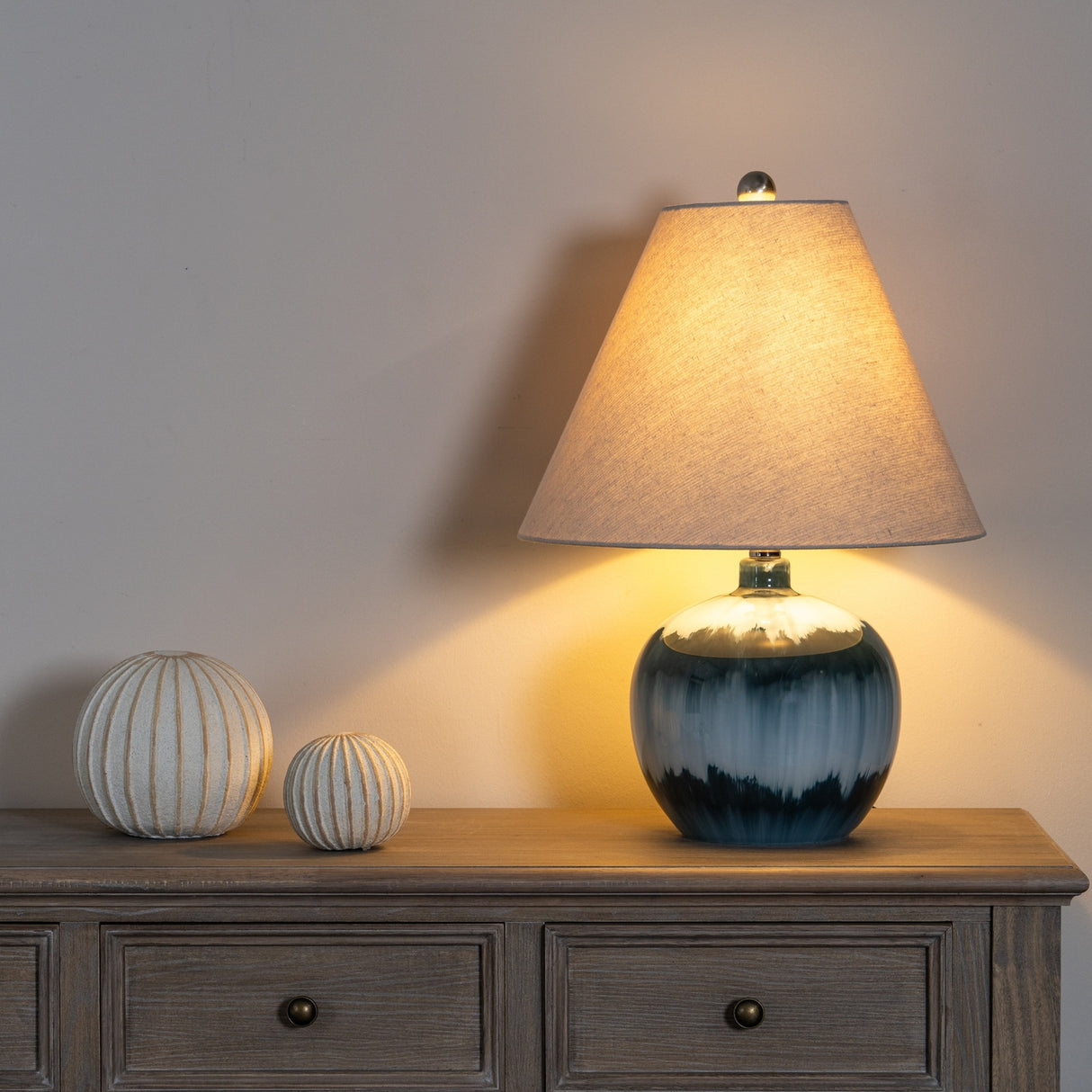 Hill Interiors Squat Glass Table Lamp in blue with rounded glass base and natural linen shade, lit on a wooden sideboard beside two ridged decorative spheres.