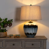 Hill Interiors Table Lamp with Linen Shade in black, glass base with drip glaze and tapered linen shade, lit on a wooden sideboard beside flowers.