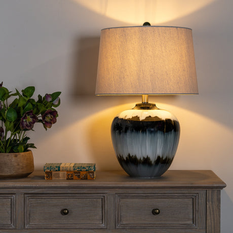 Hill Interiors Table Lamp with Linen Shade in black, glass base with drip glaze and tapered linen shade, lit on a wooden sideboard beside flowers.