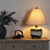 Hill Interiors Squat Glass Table Lamp with glossy black base and natural linen conical shade, lit on a wooden sideboard beside a eucalyptus vase.