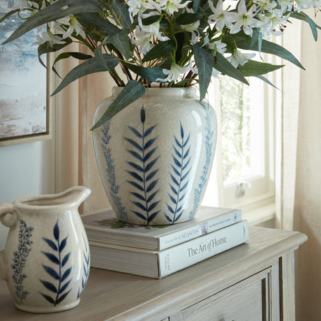 Hill Interiors Small Ceramic Olpe Vase in cream with blue leaf motifs, holding white lilies atop stacked books beside a matching jug on a console.