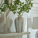 Hill Interiors Ribbed Ceramic Flora Vase in cream with blue foliage motifs, holding leafy green stems on a mantel beside candlesticks and a round mirror.