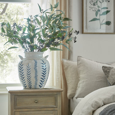 Hill Interiors Blue Flora Amphora Ceramic Vase in white with blue leaf motif and handles, holding eucalyptus and berry stems on a bedside table.