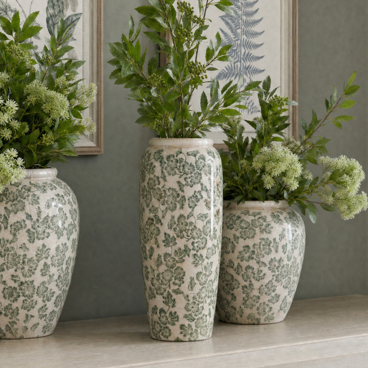 Hill Interiors Large Tall Ceramic Clover Olpe Vases in white with green floral patterning, displayed with leafy foliage on a mantel against botanical prints.