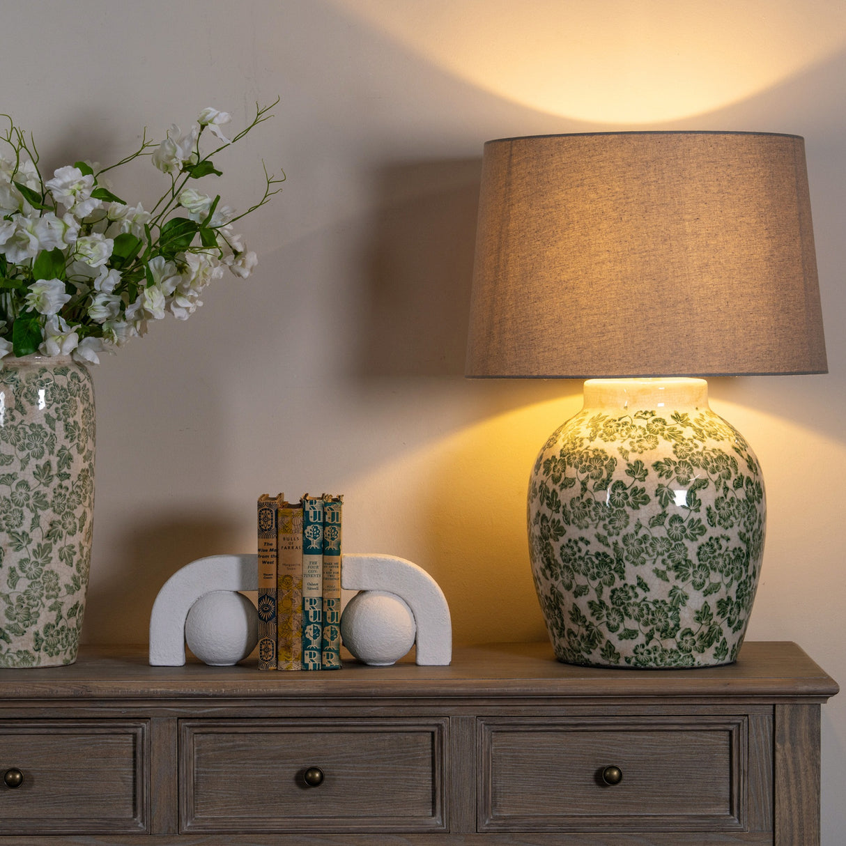 Hill Interiors Ceramic Pot Table Lamp with green floral glaze and linen shade, lit on a wooden sideboard beside a matching vase and bookends.