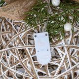 Close-up of the white LED battery unit on the Hill Interiors rattan-effect bauble, set against woven white and natural cord with foliage and berries above.