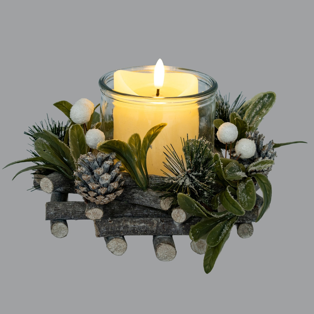 Hill Interiors Frosted Mistletoe and Pine Candle Wreath in white with pinecones and berries around a lit pillar candle in glass, on a grey background.