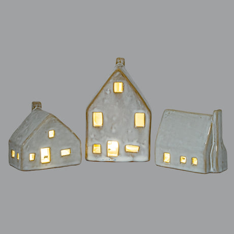 Hill Interiors Ceramic House With LED Light in white, shown as three cottage-shaped pieces of varying heights with glowing cut-out windows on a grey background.