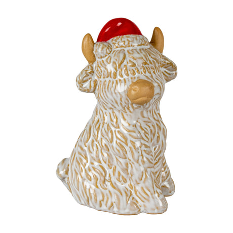 Hill Interiors ceramic Highland cow ornament in white glaze with tan horns, nose and Santa hat, seated and gazing right on a plain background.