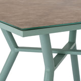 Hill Interiors French Grey Glass Table corner detail showing tempered glass top over a stone-effect surface and soft green-grey angled metal frame.