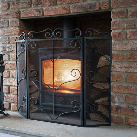Hill Interiors Heavy Large Ornate Fire Screen in black metal with scrolled detailing, set on a stone hearth before a lit fire in a brick fireplace.