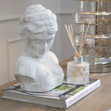 Hill Interiors Woman's Head Sculpture in cream stone with downcast gaze and styled hair, displayed on a design book beside a reed diffuser and glass lamp.