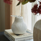 Hill Interiors Small Stone Pear ornament in beige with a matte finish and metal leaf, displayed on a stack of hardback books by a curtained window.