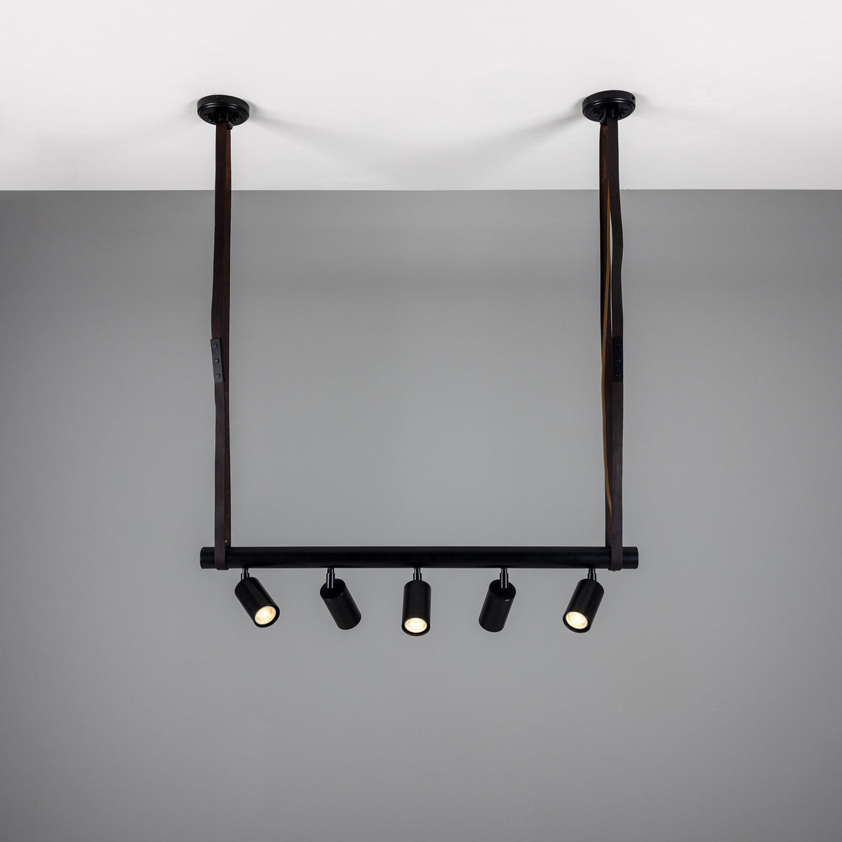 Holmes Brass 5 Light Spotlight Bar in matt black, with five cylindrical spotlights on a horizontal bar suspended by two leather straps from ceiling mounts, against a plain grey background.