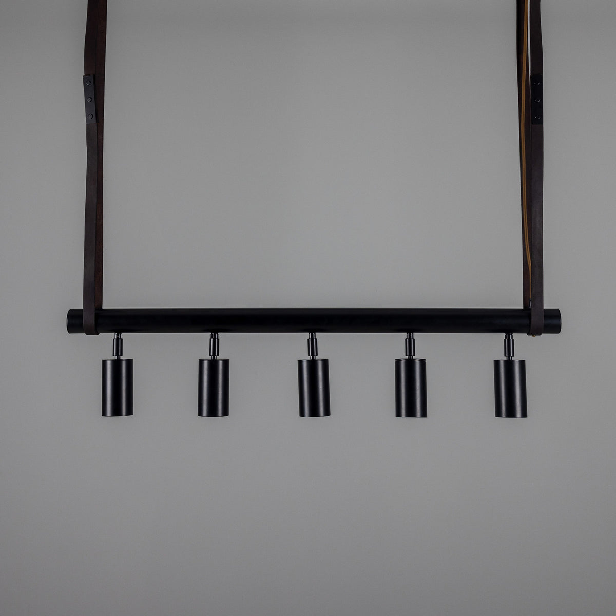 Holmes Brass 5 Light Spotlight Bar in matt black, a horizontal ceiling bar with five cylindrical spot heads, suspended by two leather straps against a plain grey background.