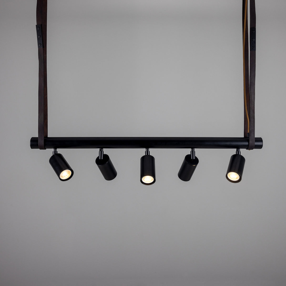 Holmes Brass 5 Light Spotlight Bar in matt black, five cylindrical spot heads on a horizontal bar, suspended by leather straps against a plain grey background.