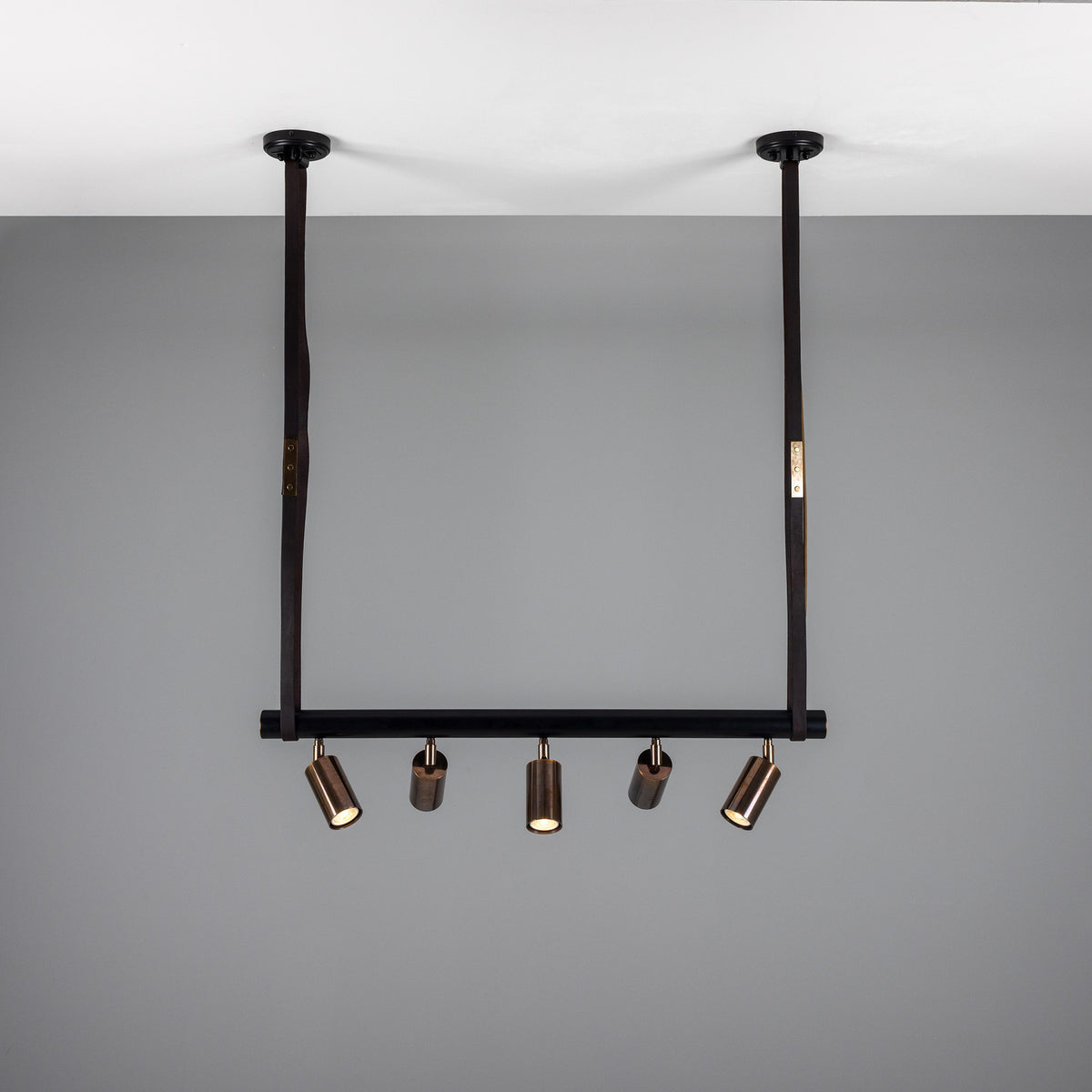 Holmes Brass 5 Light Spotlight Bar in matt black, suspended from two leather-wrapped straps with five brass cylindrical spot heads on a horizontal bar, shown against a grey ceiling and wall.