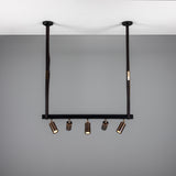 Holmes Brass 5 Light Spotlight Bar in matt black, suspended from two leather-wrapped straps with five brass cylindrical spot heads on a horizontal bar, shown against a grey ceiling and wall.