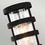 A close-up of the Hornbaek 1 Light Pedestal Light showcases its Scandinavian design with a cylindrical shape. The black structure features clear glass encasing a glowing filament bulb, prominently displayed against a neutral background. This pedestal light combines elegance and functionality in any setting.