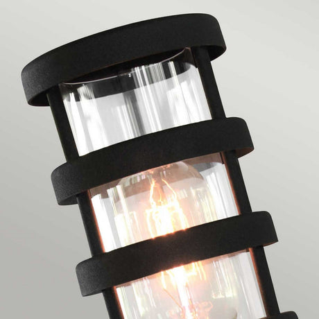 A close-up of the Hornbaek 1 Light Pedestal Light showcases its Scandinavian design with a cylindrical shape. The black structure features clear glass encasing a glowing filament bulb, prominently displayed against a neutral background. This pedestal light combines elegance and functionality in any setting.