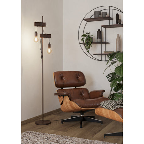 Hosmane Floor Lamp in antique brown steel, slim pole with round base, two staggered girder brackets holding hanging E27 bulbs, shown beside a leather lounge chair.