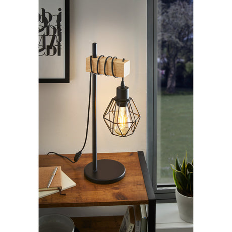 Hosmane black steel table lamp with wooden crossbar and geometric wire cage shade, lit on a wooden desk beside a window.