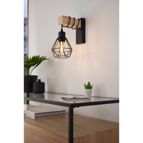 Hosmane black wall light with wooden arm and wire-cage shade, cable looped around the timber, shown lit above a glass-top console table in a living room.