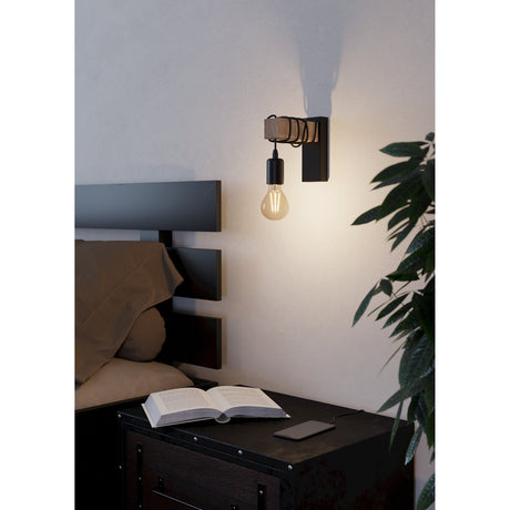 Hosmane Wall Light with black backplate and pale wood beam, black cord looped along the arm to a bare E27 bulb, shown above a bedside table in a bedroom setting.