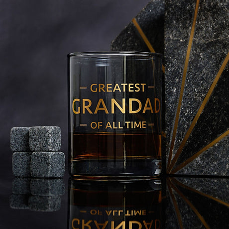 Straight-sided glass whiskey tumbler with a smoky blue tint, gold "GREATEST GRANDAD OF ALL TIME" text, shown with four grey whiskey stones on a dark reflective surface.