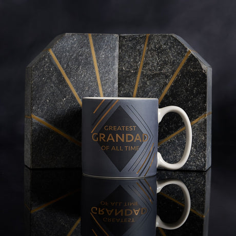 Matt blue mug with "Greatest Grandad of All Time" in gold text inside a charcoal diamond, white handle, displayed against a dark geometric marble backdrop.