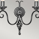 Close-up of the Harlech 2 Light Wall Light in black, showcasing two candelabra-style arms on either side. It features a central round base and a medieval-inspired curled design hanging below. The plain light gray background highlights the sconce's intricate details and elegant black finish.